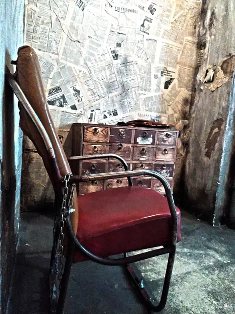 Fauteuil enchaîné devant un mur couvert de journaux et un vieux meuble à tiroirs
